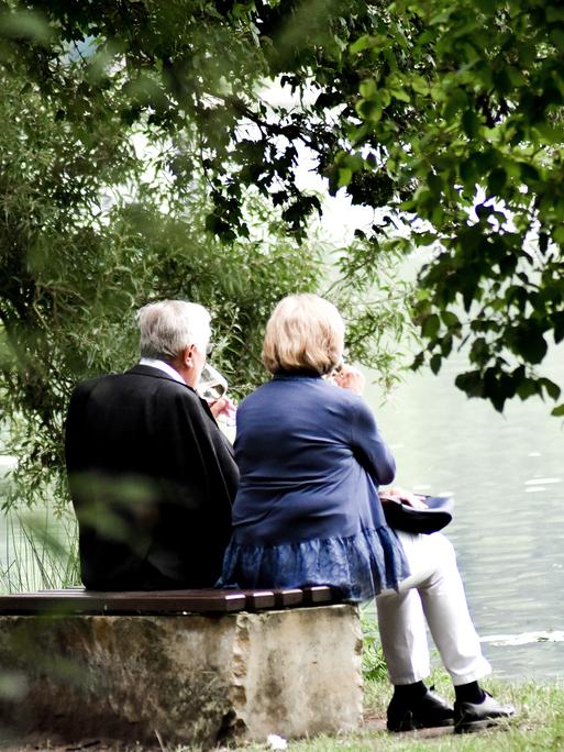 Ein älteres Ehepaar sitzt auf einer Bank vor einem See und trinkt Wein.