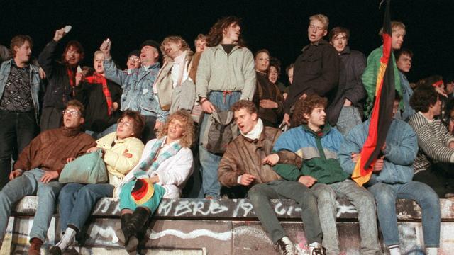 Ein Foto von 1989: junge Menschen sitzen in Berlin auf der Mauer. Sie schunkeln und freuen sich. 