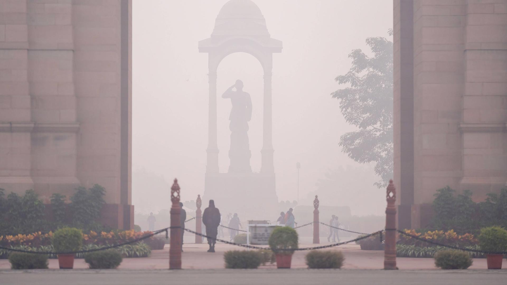 Eine Statue in der Stadt Neu Delhi, davor laufen einige Menschen. Alles liegt unter einer dicken Smog-Schicht.