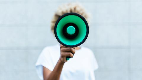 Vorderansicht einer Frau mit Afro-Frisur, deren Gesicht durch ein Megafon verdeckt wird, das sie frontal in die Kamera hält.