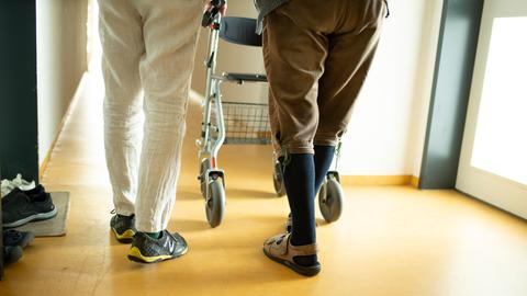 Eine Person begleitet einen Mann mit Rollator im Hausflur. Es sind vor allem die Beine und Füsse zu sehen.