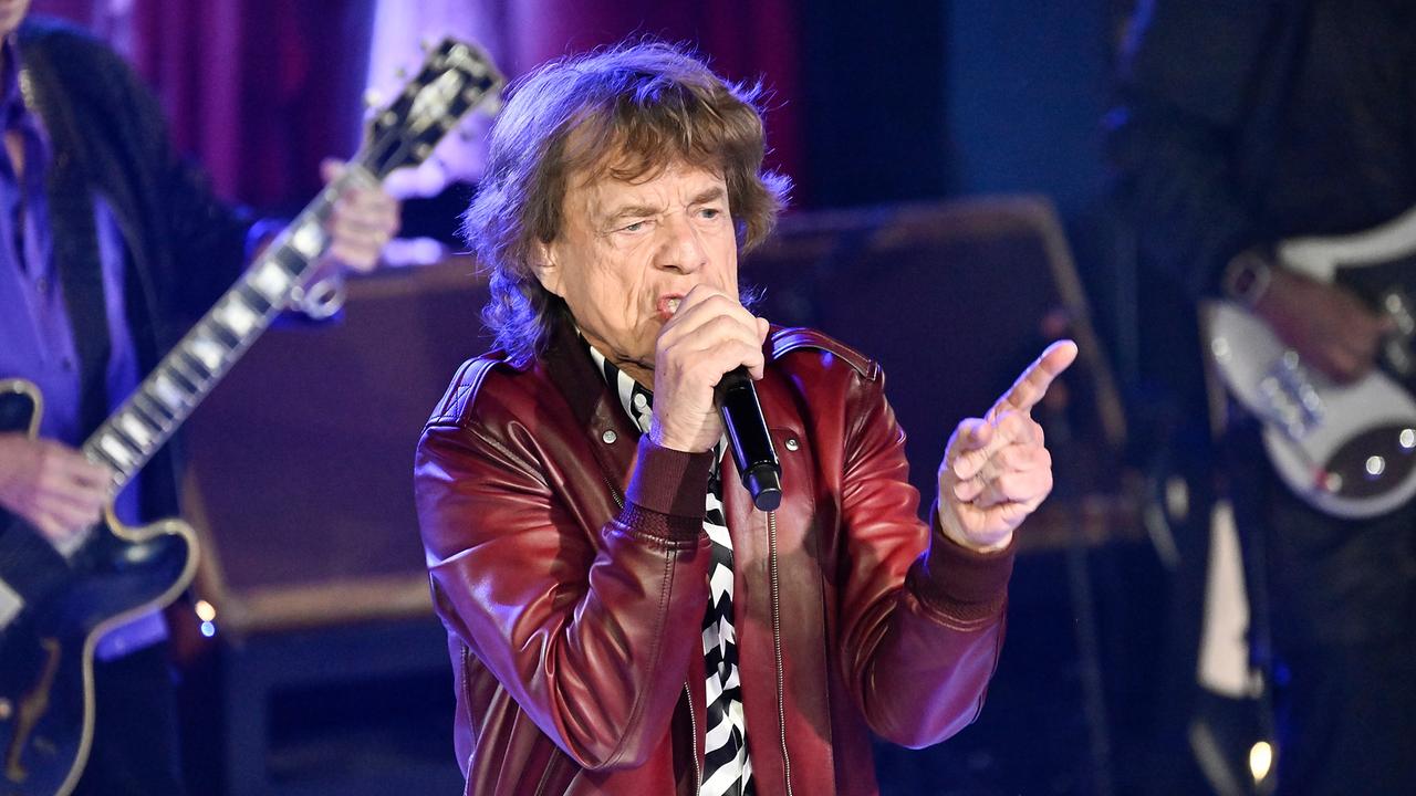  Mick Jagger in einer rötlichen Lederjacke singt auf einer Bühne. Daneben sieht man die Gitarren seiner Bandkollegen.