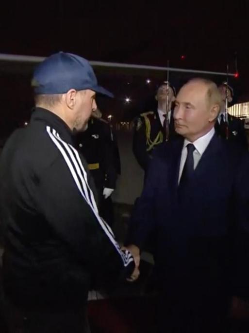 Wladmir Putin begrüßt auf dem nächtlichen Moskauer Flughafen einen Mann im Trainingsanzug mit Basecap per Handschlag.