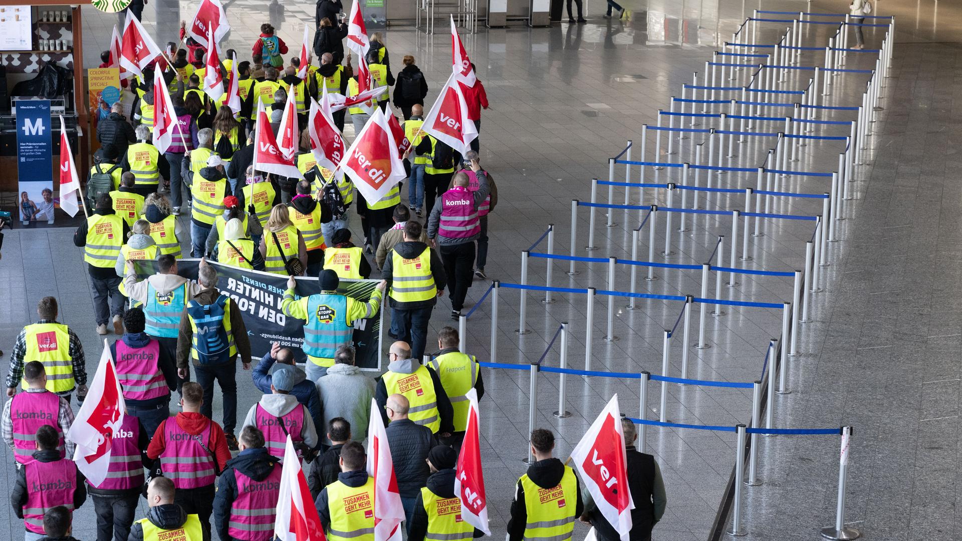Verdi-Mitglieder ziehen in einem Protestzug durch ein Terminal im Flughafen. Sie sind von oben fotografiert; man sieht sie von hinten. Verdi-Mitglieder ziehen in einem Protestzug durch ein Terminal im Flughafen. Sie sind von oben fotografiert; man sieht sie von hinten.