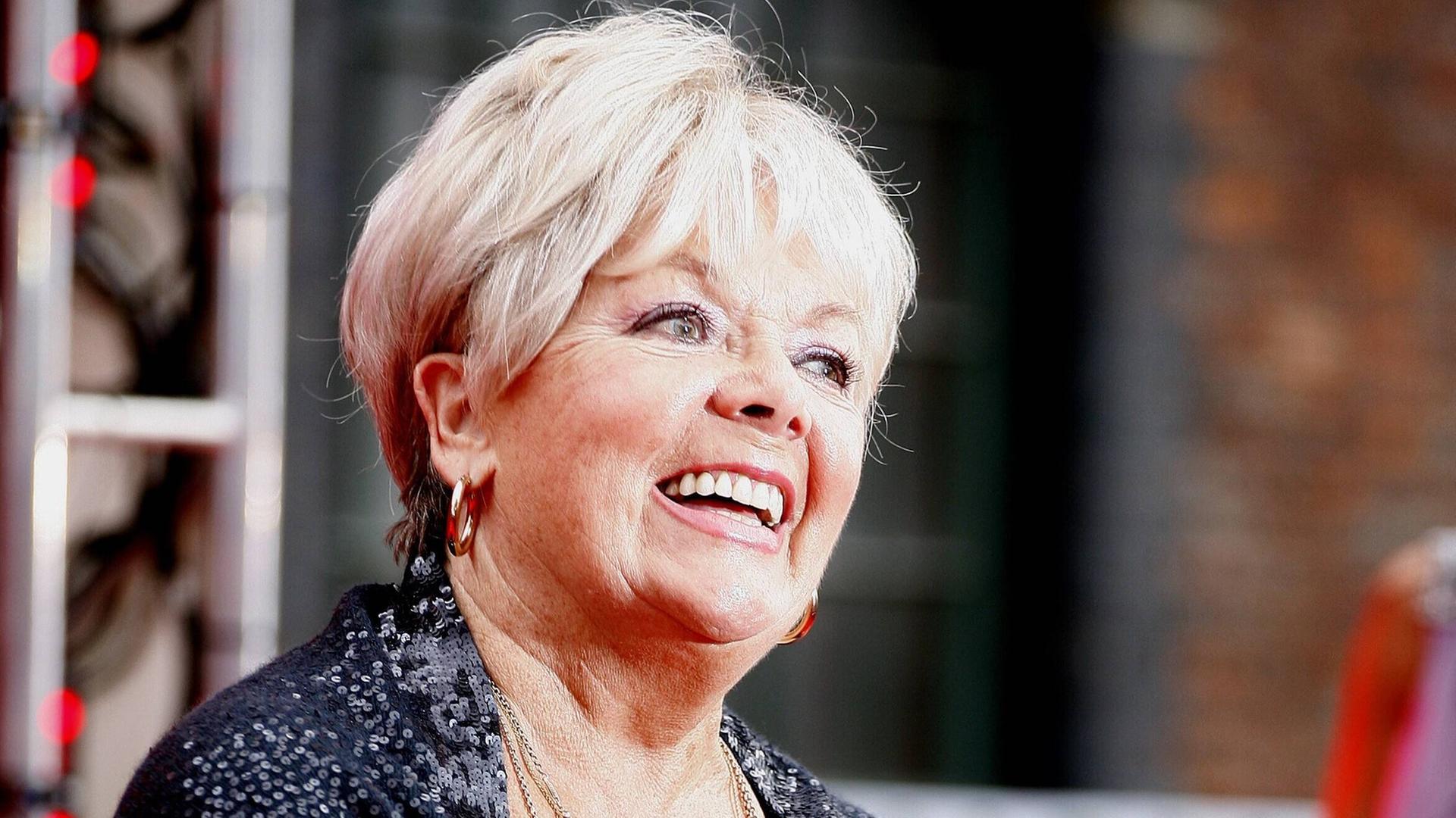 Die britische Schauspielerin Meg Johnson lächelt in einem schwarzen Umhang in die Kamera.