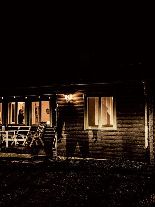 Es ist Nacht, im Holzhaus brennt Licht, auf der Fassade sieht man den großen Schatten eines Mannes.