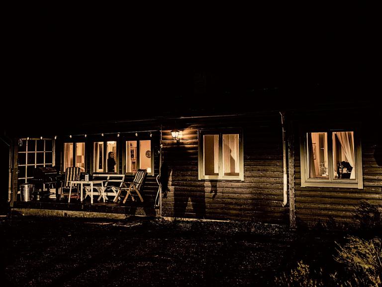 Es ist Nacht, im Holzhaus brennt Licht, auf der Fassade sieht man den großen Schatten eines Mannes.