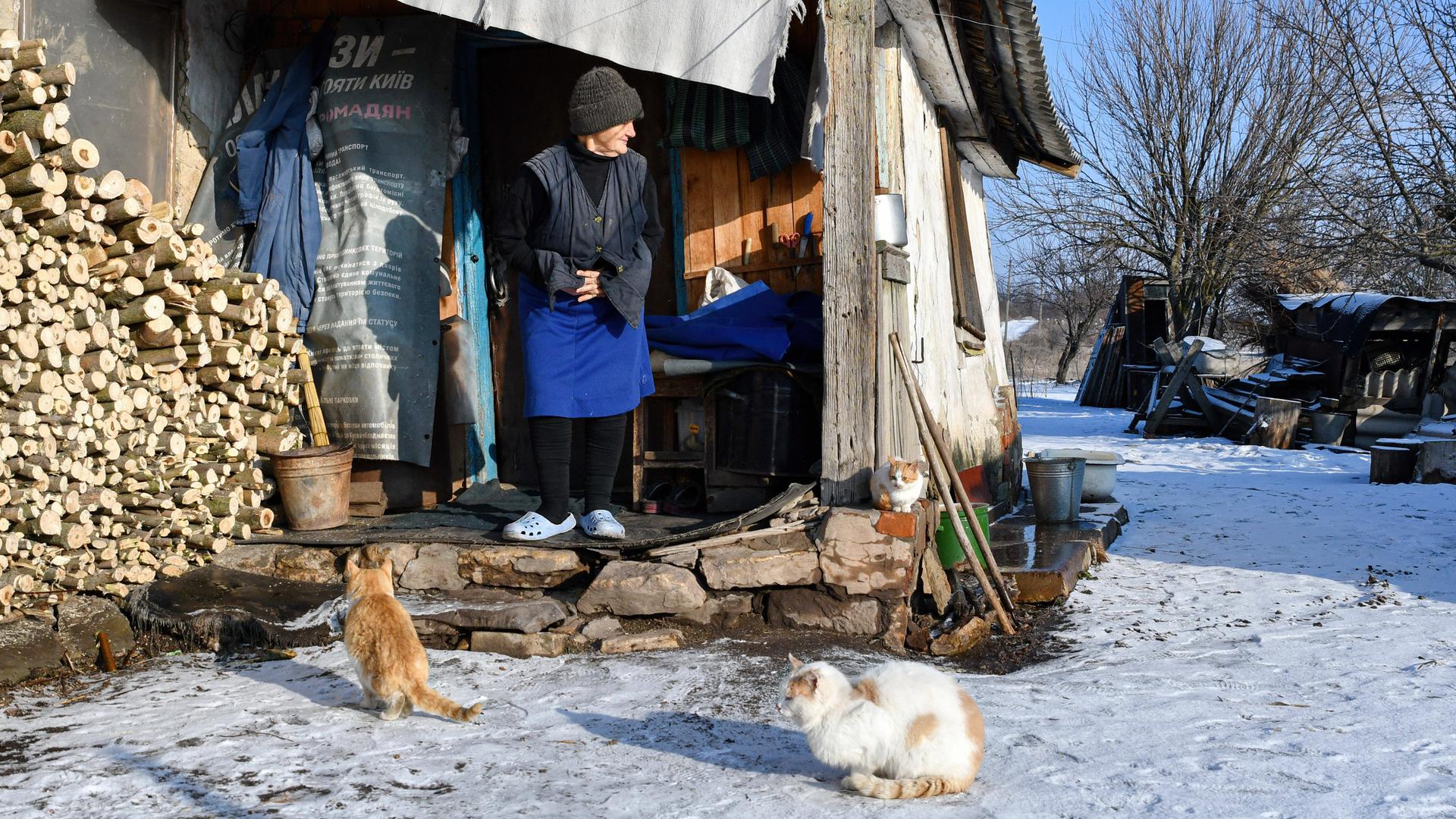 Ukraine: Sofiya (85 jahre alt) steht vor Ihrem Haus nahe der Frontlinie. Im Schnee vor Ihr sitzen zwei Katzen. 5.Februar 2022, Zaitseve, Donetsk, Ukraine.