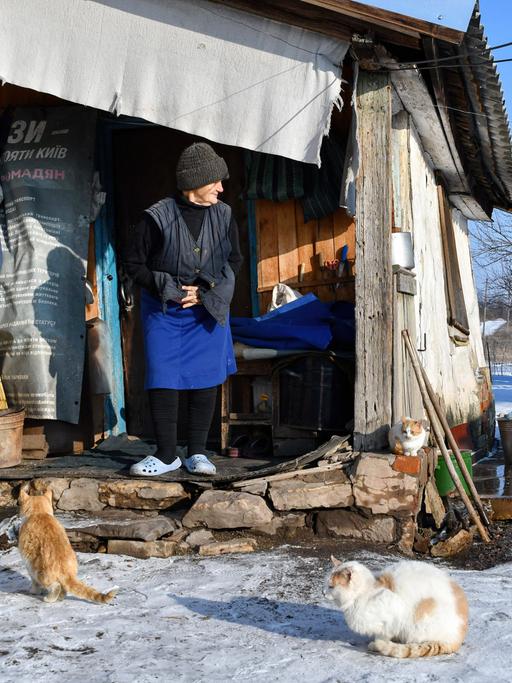 Ukraine: Sofiya (85 jahre alt) steht vor Ihrem Haus nahe der Frontlinie. Im Schnee vor Ihr sitzen zwei Katzen. 5.Februar 2022, Zaitseve, Donetsk, Ukraine.