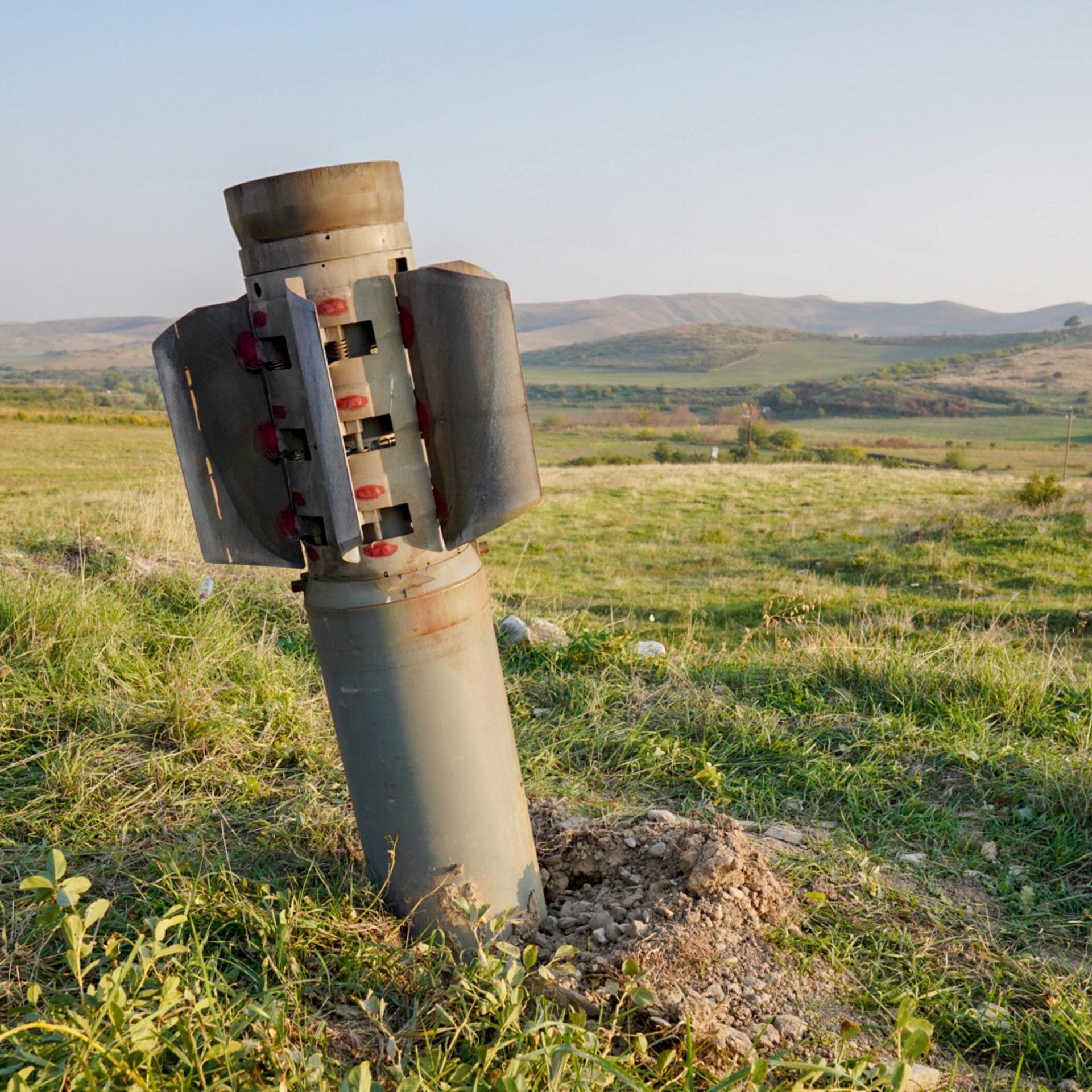Ein Streubomben-Geschoss steckt im Boden, im Hintergrund ein hügelige Graslandschaft