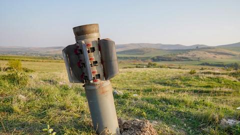 Ein Streubomben-Geschoss steckt im Boden, im Hintergrund ein hügelige Graslandschaft