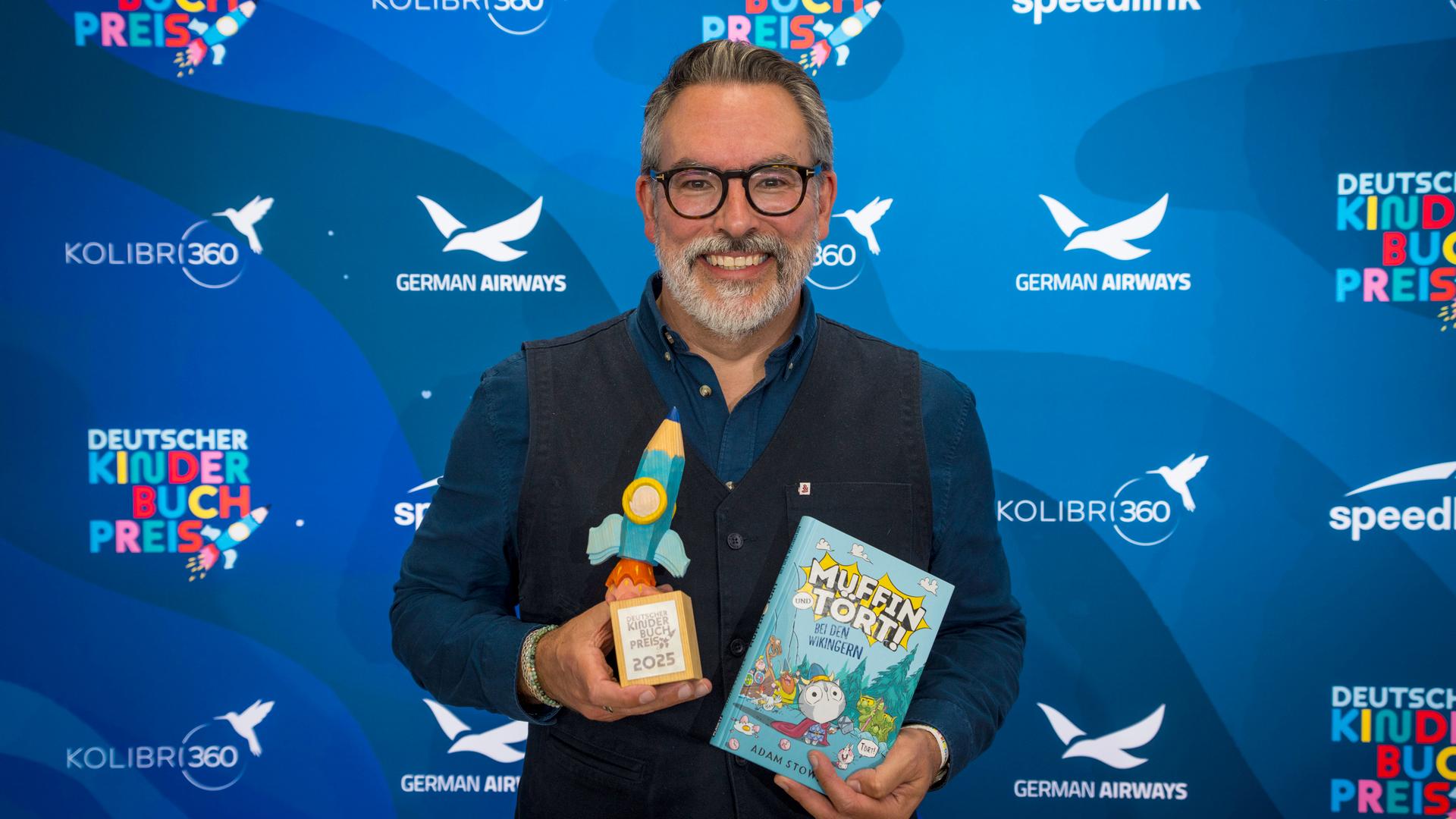 Der Autor Adam Stower hält den deutschen Kinderbuchpreis in einer Hand, in der anderen sein Buch "Muffin und Tört". Der Autor Adam Stower hält den deutschen Kinderbuchpreis in einer Hand, in der anderen sein Buch "Muffin und Tört".