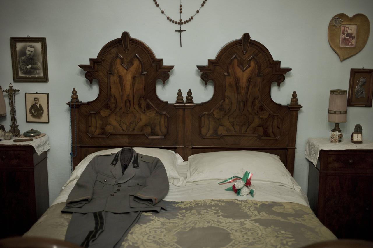 Ein Doppelbett. Auf der einen Seite liegt ein gebügelter Anzug mit Abzeichen. An den Wänden Fotos und ein Kreuz.