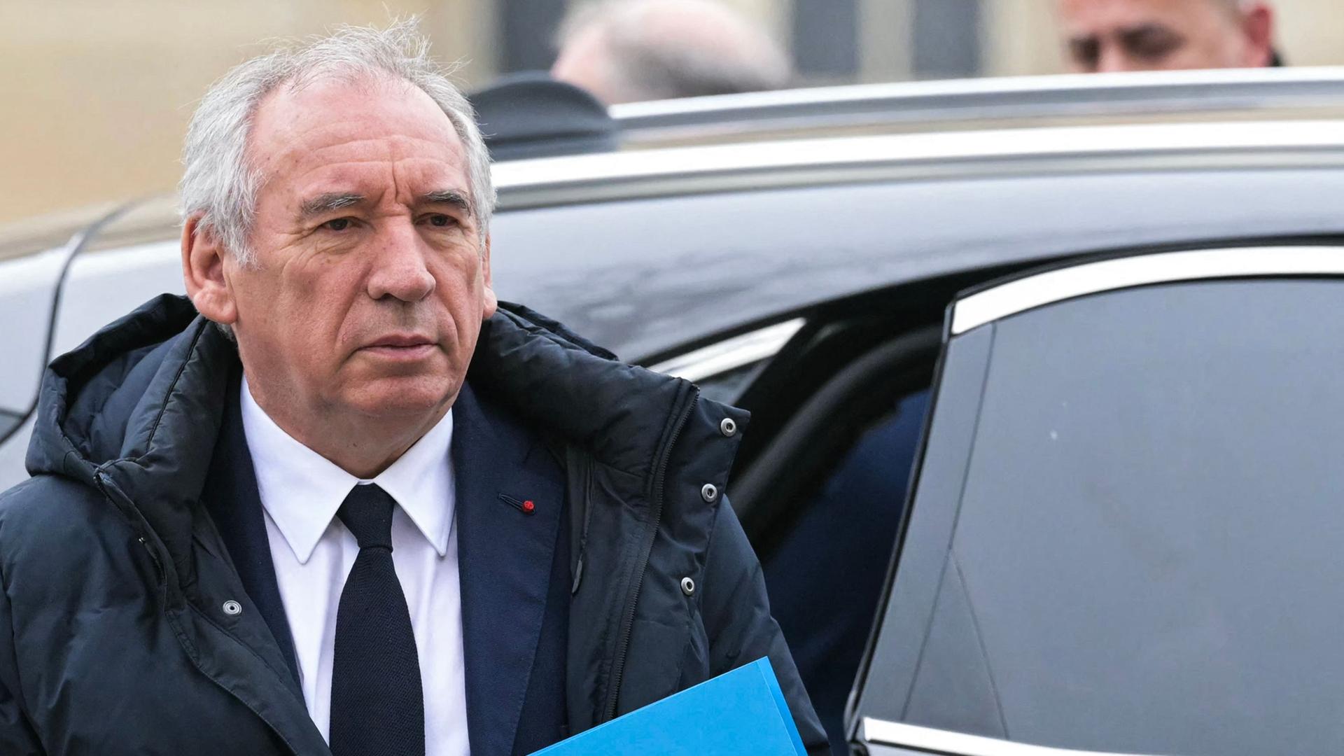 Bayrou in einer schwarzen Jacke und mit einem blauen Ordner in der Hand steht vor der geöffneten Tür eines Autos. Er blickt ernst. 