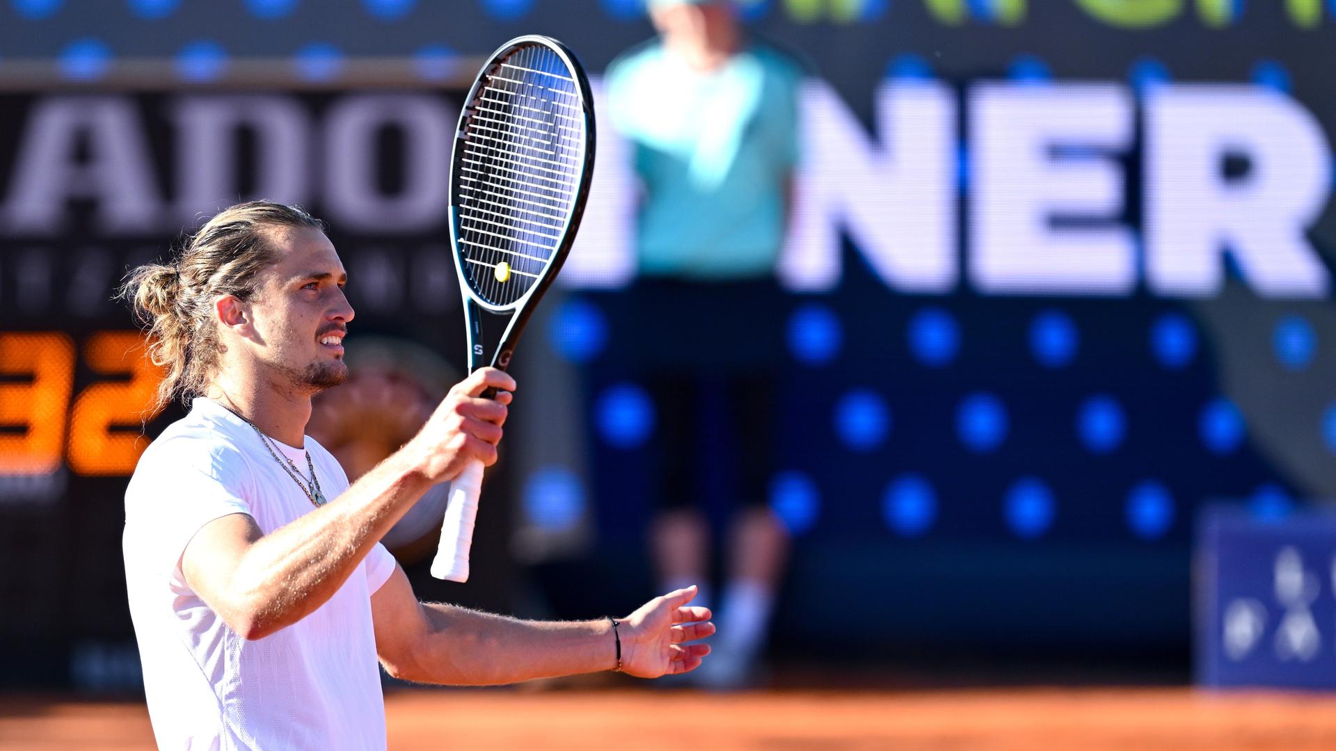 Alexander Zverev hält den Tennisschläger mit der linken Hand hoch. Er steht auf dem Tennisplatz und trägt ein weißes Shirt. Er lächelt. Alexander Zverev hält den Tennisschläger mit der linken Hand hoch. Er steht auf dem Tennisplatz und trägt ein weißes Shirt. Er lächelt.