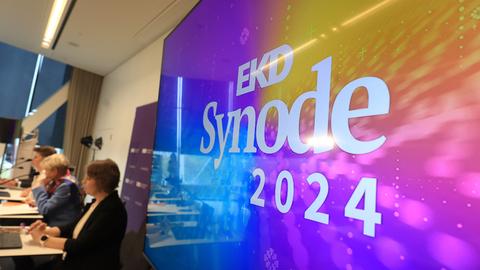 Würzburg: Mitglieder des Präsidiums sitzt bei der Synode der Evangelischen Kirche in Deutschland (EKD) bei einer Pressekonferenz. Im Bildvordergrund das Logo der Synode auf einem großen Bildschirm.