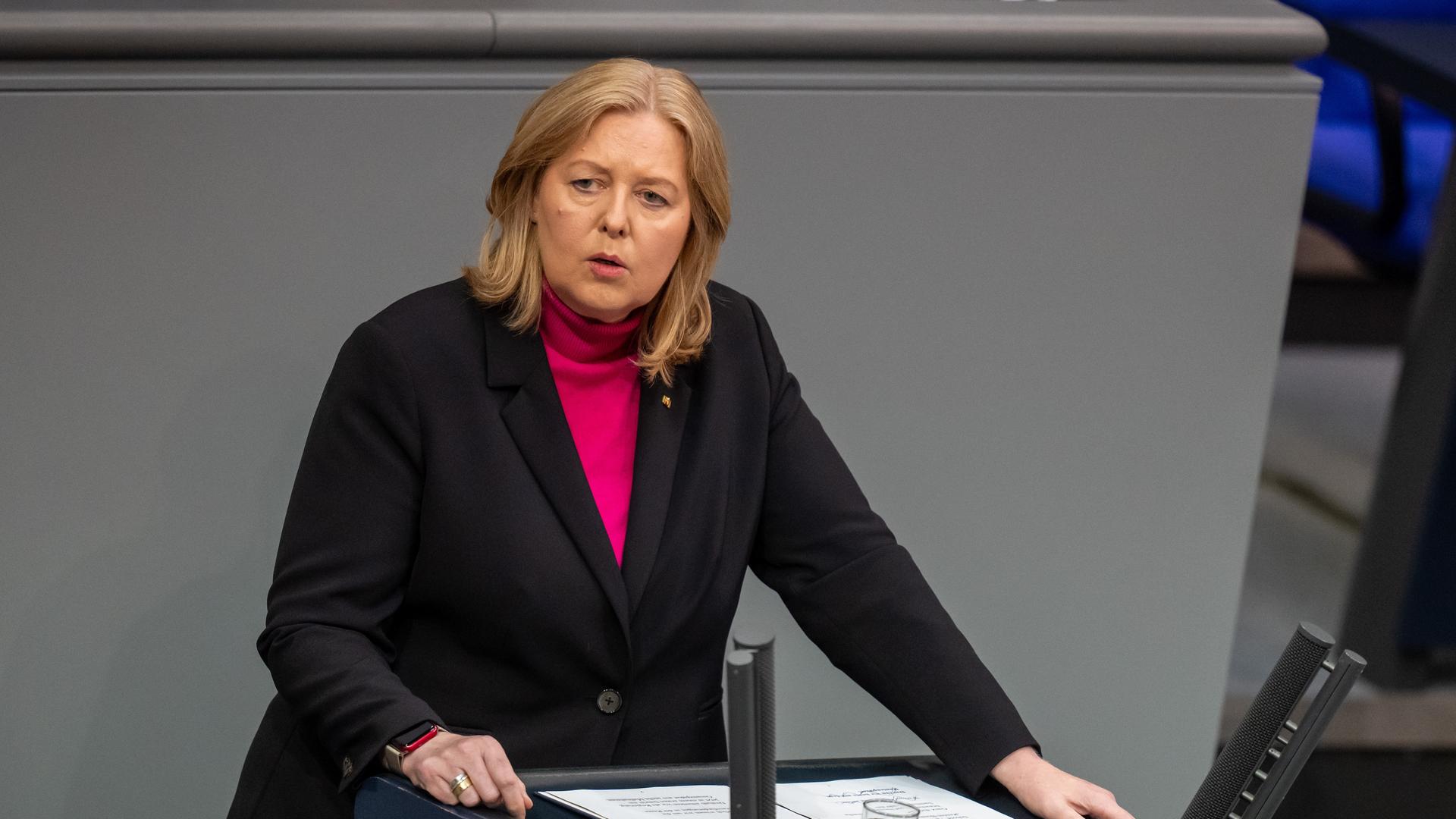 Bas steht am Rednepult und sagt etwas. Sie trägt ein schwarzes Jackett über einer magentafarbenen Bluse. 