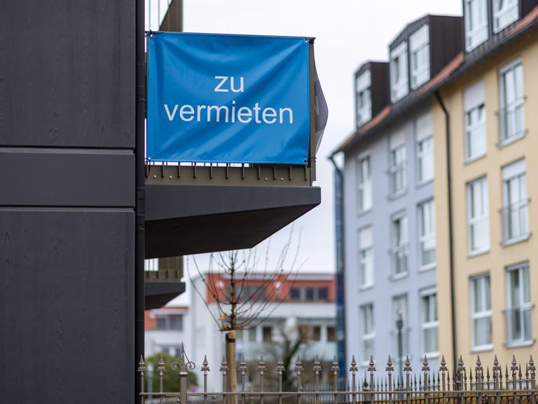 "Zu vermieten" steht auf einer Plane, die an einem Balkon-Geländer einer Neubau-Wohnung angebracht ist.
