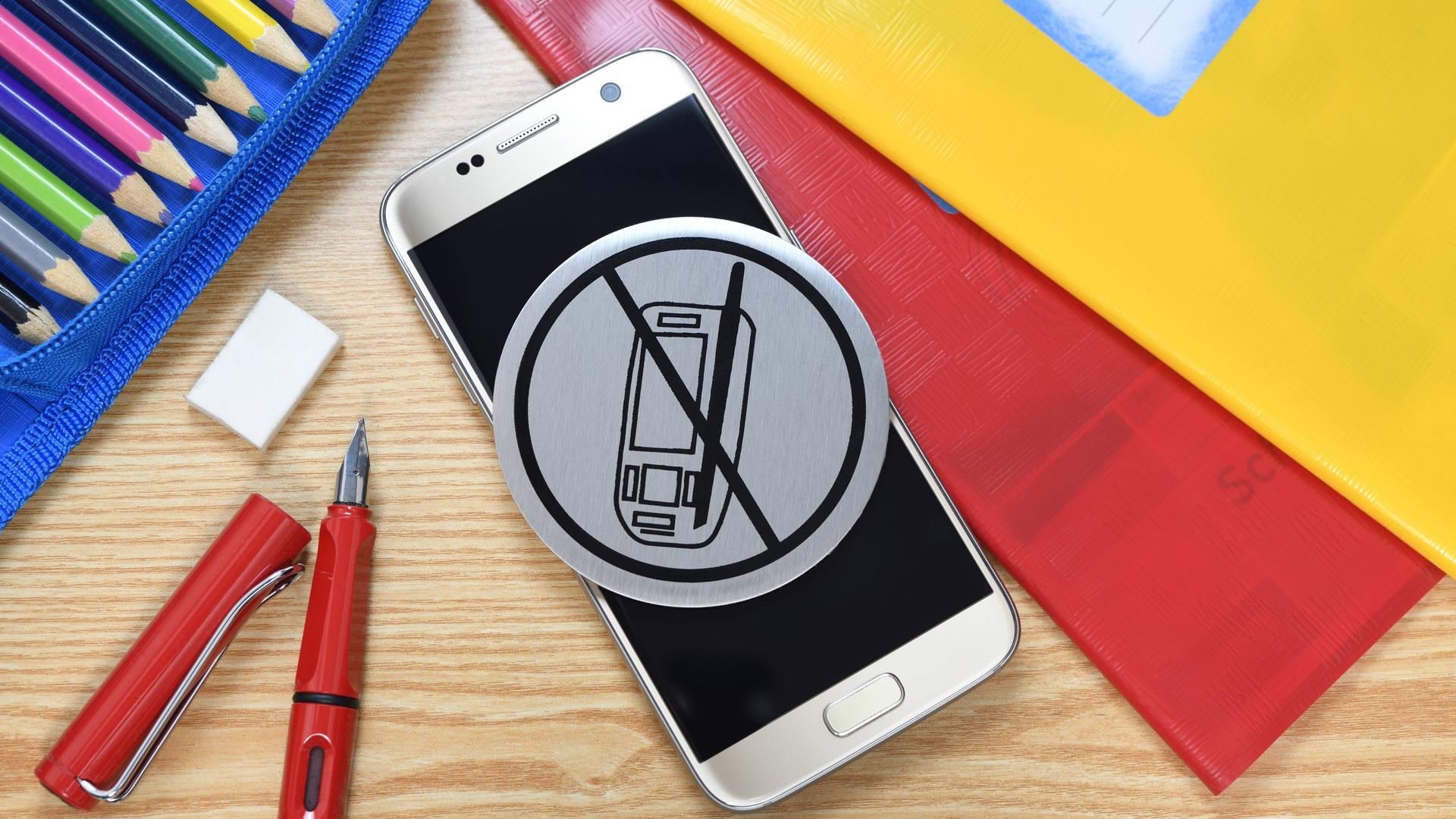 Ein Handy mit Verbotsschild umgeben von Schulutensilien Ein Handy mit Verbotsschild umgeben von Schulutensilien