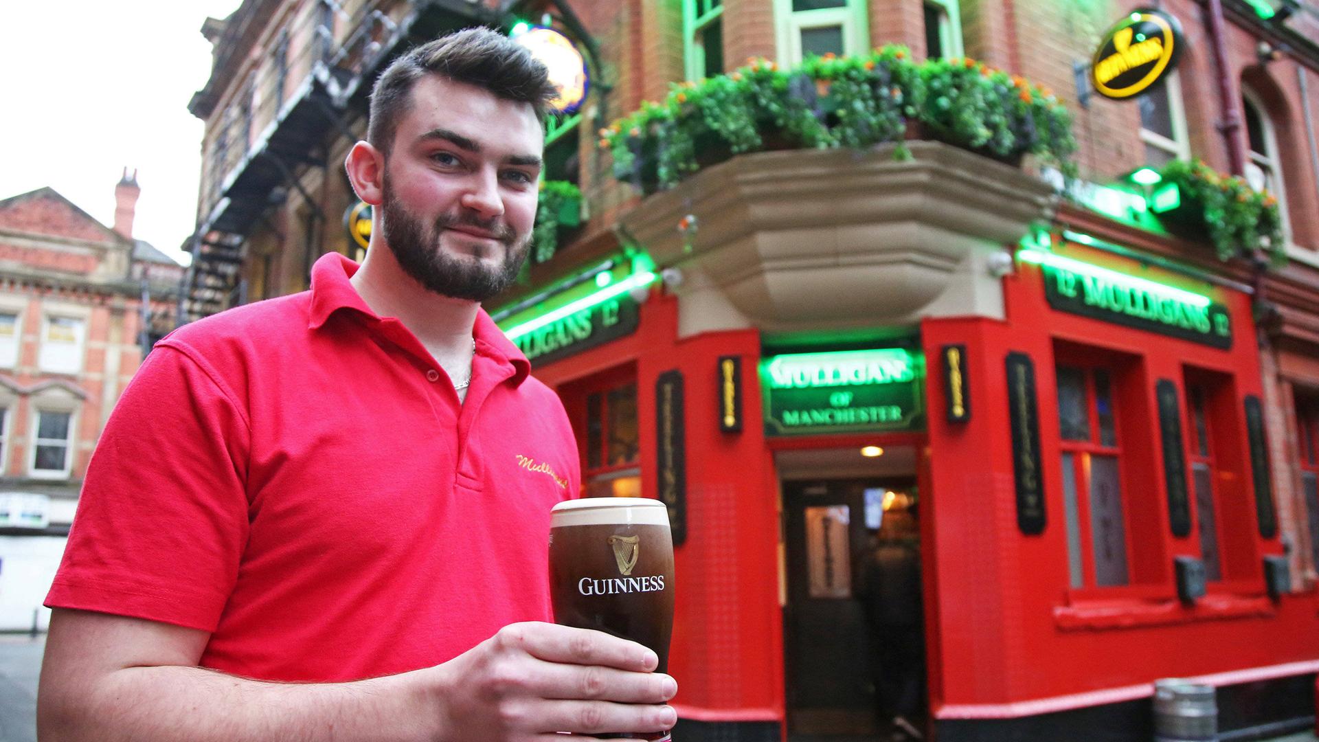 Der Mann mit schwarzem Bart und rotem Shirt steht mit dem Glas in der Hand vor der Bar mit roten Wänden und lächelt in die Kamera.