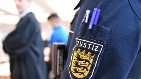 Ein Angeklagter steht im Juli 2020 im Gerichtssaal in der Messe Ulm, während im Vordergrund eine Justizmitarbeiterin steht, deren Justiz-Logo am Ärmel zu sehen ist. Fünf Männer sind im Juli 2020 wegen Vergewaltigung einer 14-Jährigen angeklagt.