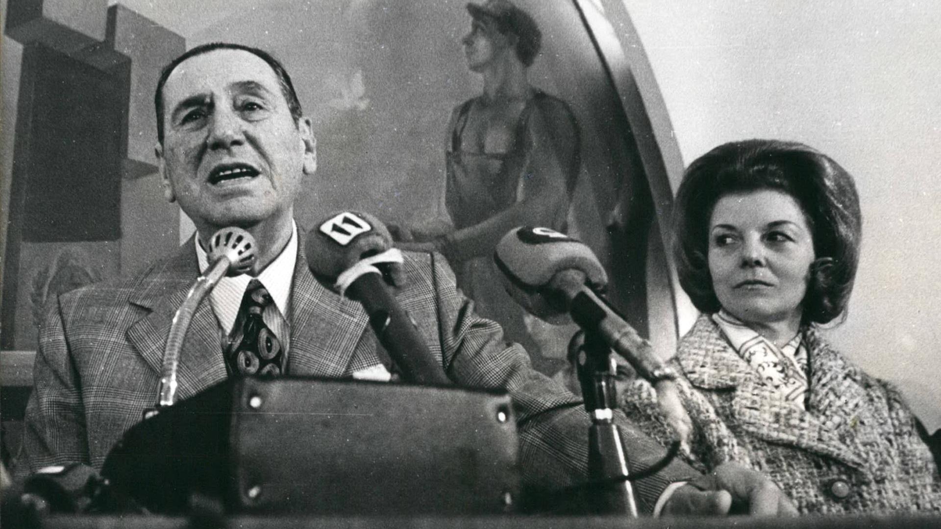 Juan Domingo Perón steht neben seiner Frau an einem Rednerpult und spricht engagiert ins Mikrofon