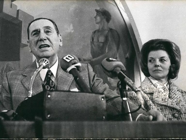 Juan Domingo Perón steht neben seiner Frau an einem Rednerpult und spricht engagiert ins Mikrofon