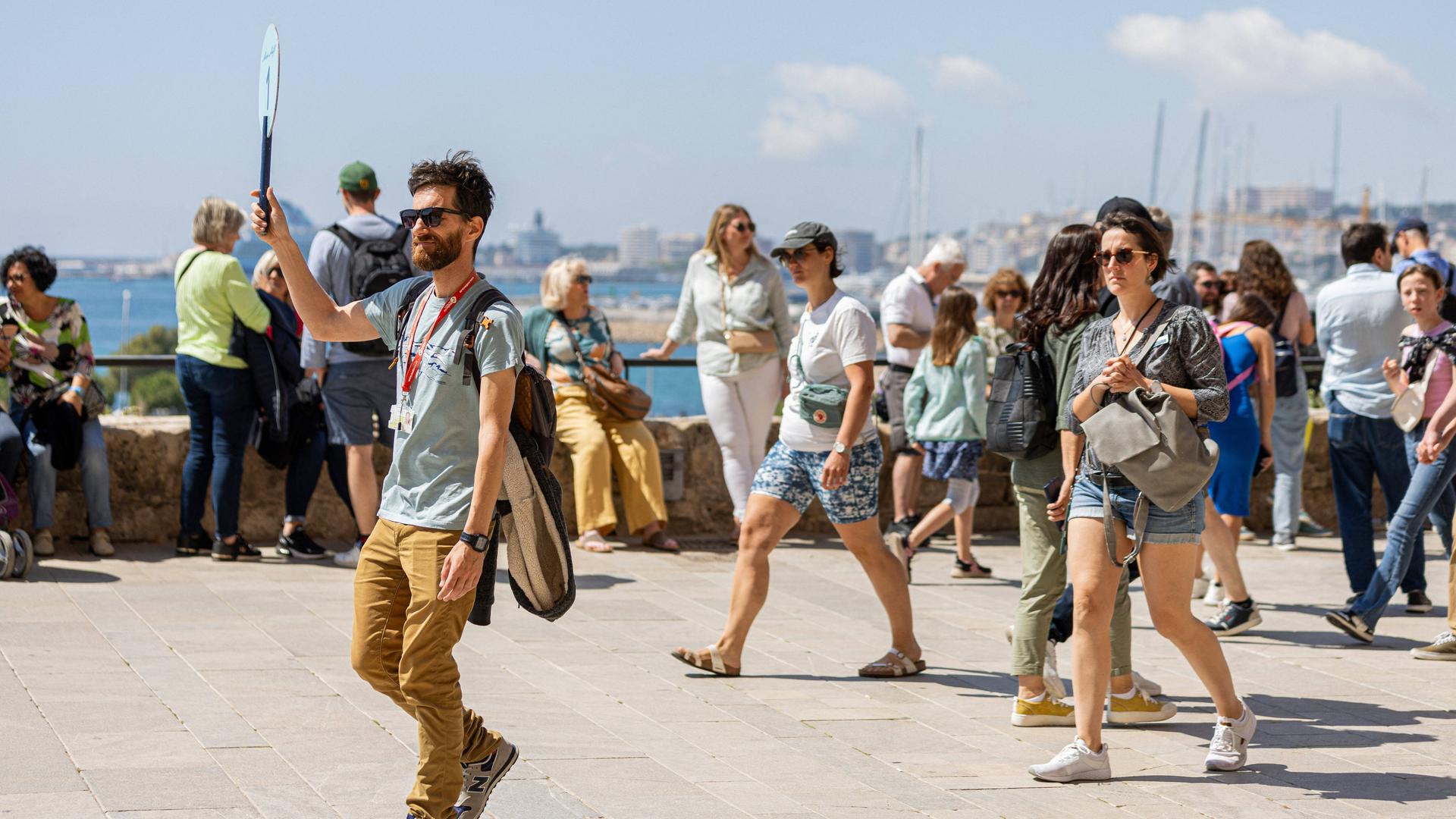 Ein Touristenführer in Mallorca hält ein Schild hoch. Hinter ihm sind weitere Menschen auf der Straße zu sehen, im Hintergrund ist das Meer zu erkennen.