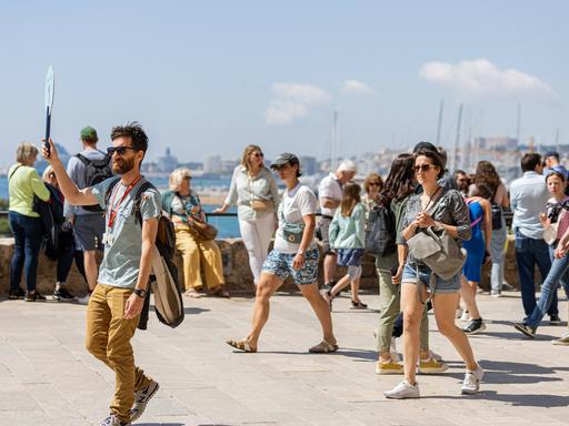 Ein Touristenführer in Mallorca hält ein Schild hoch. Hinter ihm sind weitere Menschen auf der Straße zu sehen, im Hintergrund ist das Meer zu erkennen.