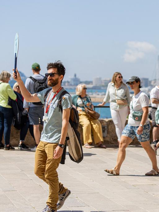 Ein Touristenführer in Mallorca hält ein Schild hoch. Hinter ihm sind weitere Menschen auf der Straße zu sehen, im Hintergrund ist das Meer zu erkennen.