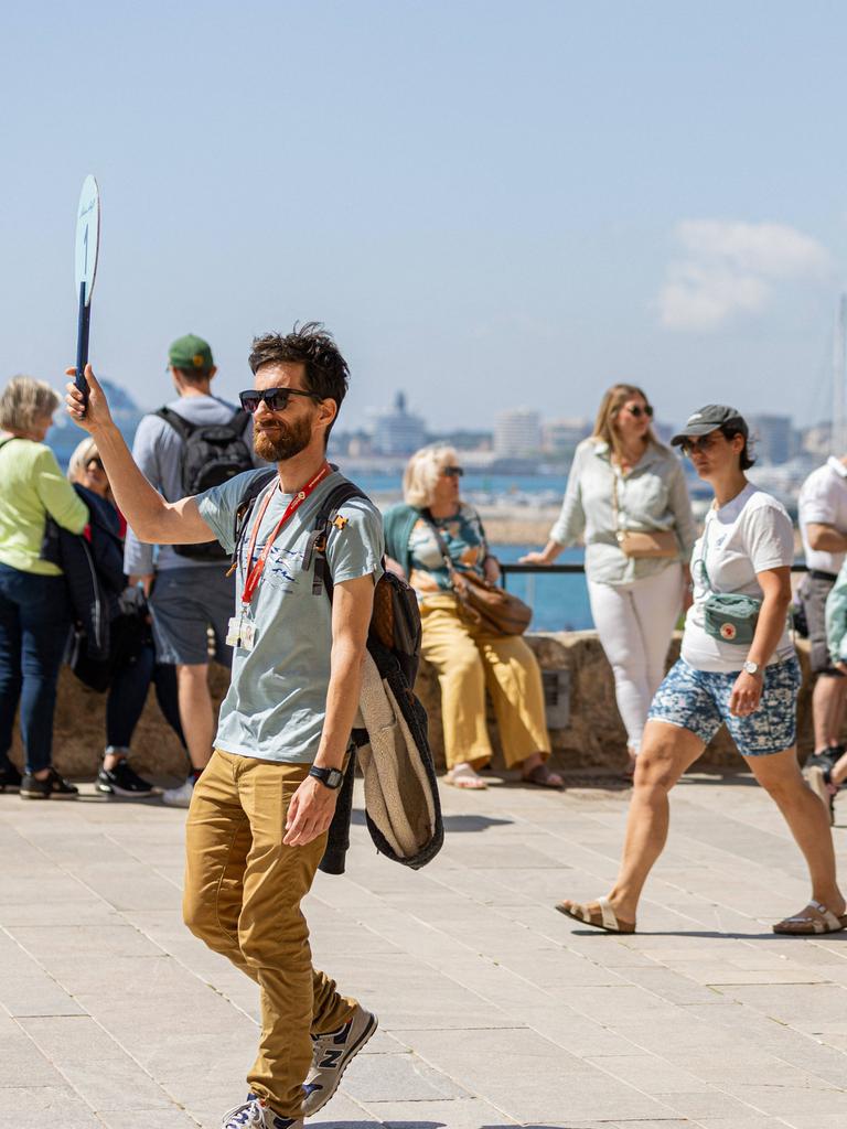 Ein Touristenführer in Mallorca hält ein Schild hoch. Hinter ihm sind weitere Menschen auf der Straße zu sehen, im Hintergrund ist das Meer zu erkennen.