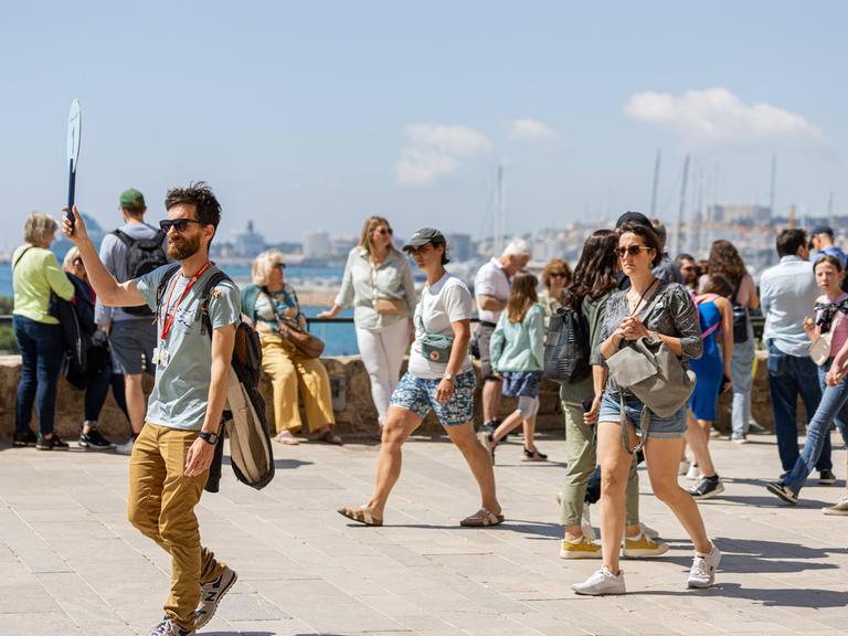 Ein Touristenführer in Mallorca hält ein Schild hoch. Hinter ihm sind weitere Menschen auf der Straße zu sehen, im Hintergrund ist das Meer zu erkennen.
