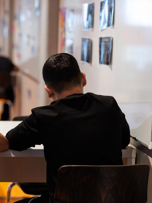 Schüler einer Realschule in München schreiben während einer Unterrichtsstunde in ihre Schulhefte.Man sieht sie von hinten mit schwarzen T-Shirts am Tisch sitzend. 