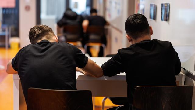 Schüler einer Realschule in München schreiben während einer Unterrichtsstunde in ihre Schulhefte.Man sieht sie von hinten mit schwarzen T-Shirts am Tisch sitzend. 