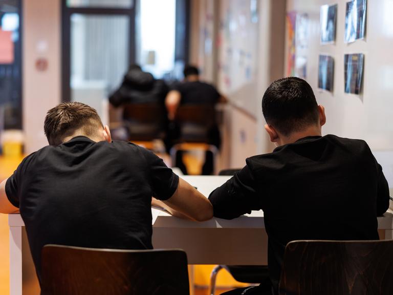 Schüler einer Realschule in München schreiben während einer Unterrichtsstunde in ihre Schulhefte.Man sieht sie von hinten mit schwarzen T-Shirts am Tisch sitzend. 