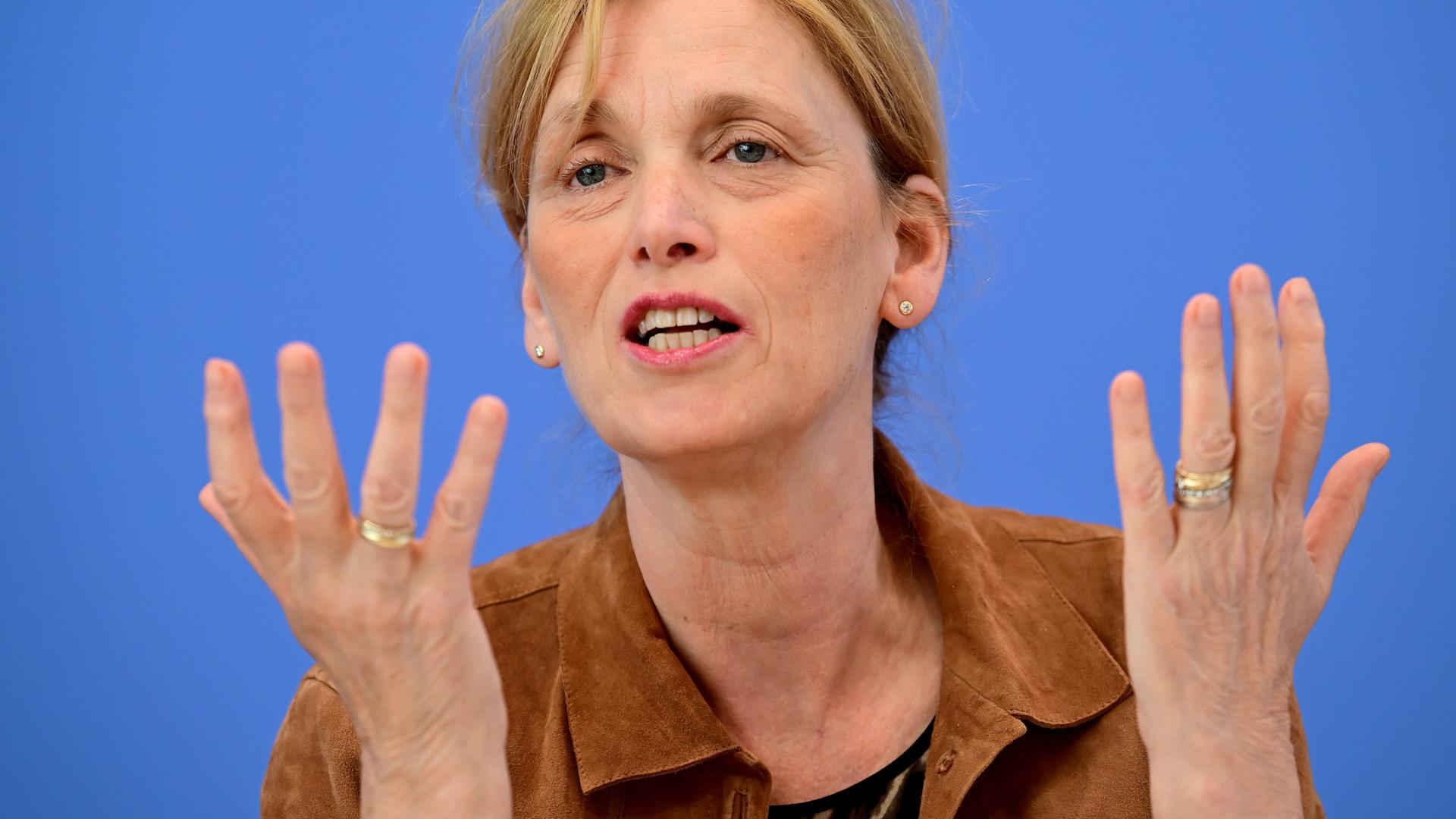 Karin Prien sitzt vor einem blauen Hintergrund und spricht gestikulierend.