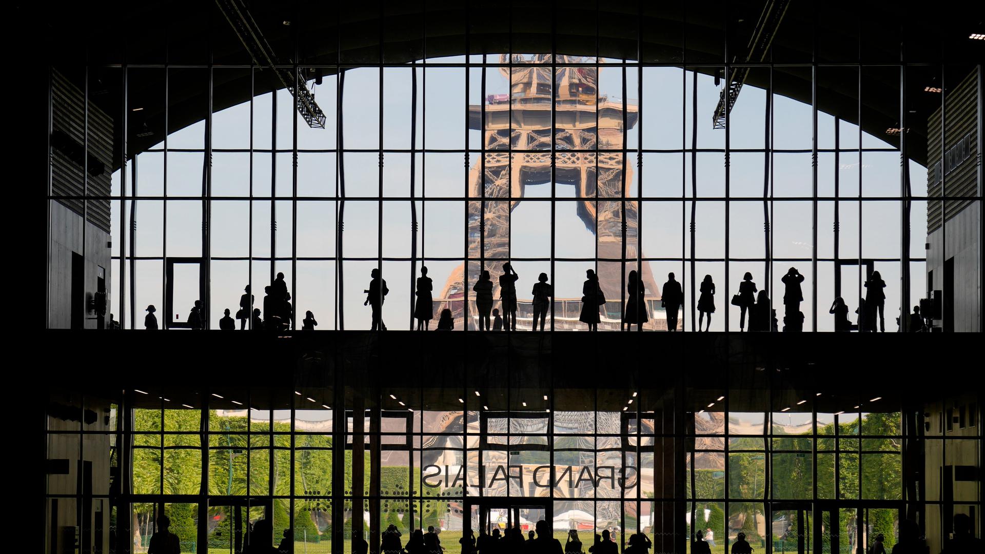 Eine Fensterwand des Grand Palais mit Besuchern als Silhouetten auf zwei Ebenen. Hinter des Fensterwand sieht man den Eiffelturm. 