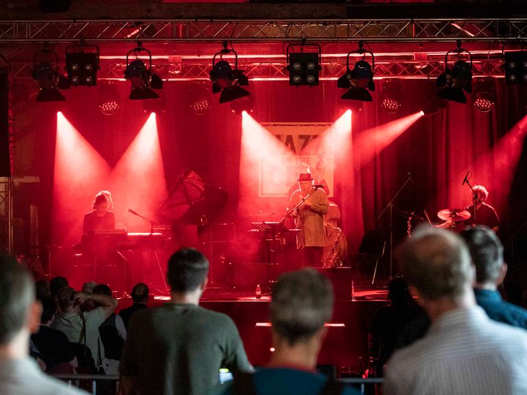 Konzert von Max Herre & Roberto di Gioia live auf dem Jazz in the City Festivals im Zughafen Erfurt. Man sieht die Zuschauer, die auf die Bühne blicken. Auf der Bühne stehen in rotes Licht getaucht die Musiker.