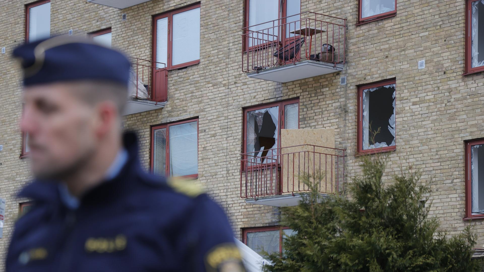 Ein schwedischer Polizist (verschwommen) blickt nach links, Hintergrund ein Wohnhaus mit einer zersprungenen Fensterscheibe