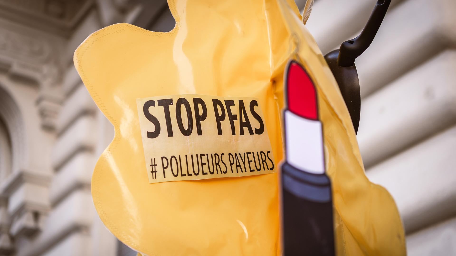 Ein Aufkleber mit der Aufschrift "Stop PFAS" und einer Abbildung eines Lippenstifts sind auf einem aufblasbaren, gelben Plastikballon befestigt.