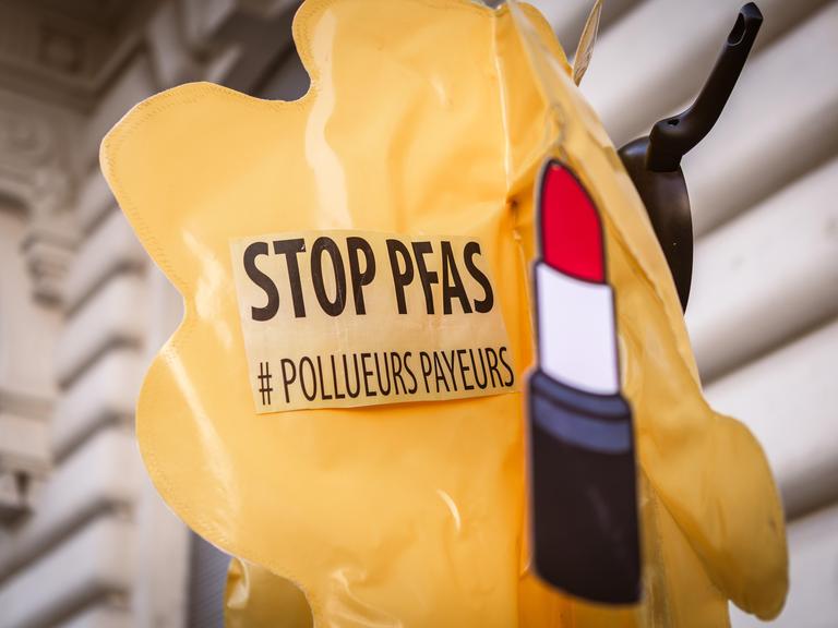 Ein Aufkleber mit der Aufschrift "Stop PFAS" und einer Abbildung eines Lippenstifts sind auf einem aufblasbaren, gelben Plastikballon befestigt.