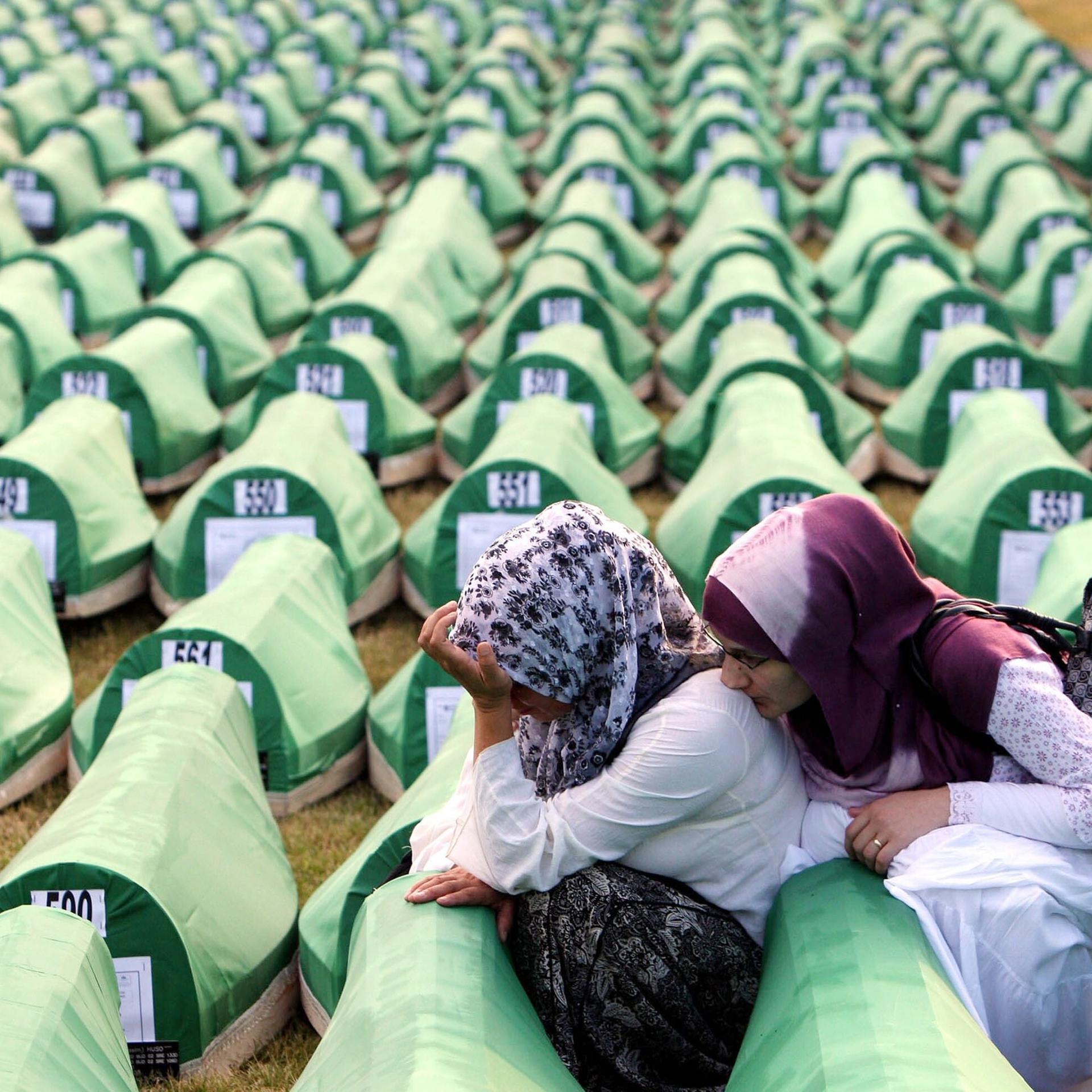 Zwei Frauen trauern während der Beerdigung von neu identifizierten bosnischen Muslimen in Srebrenica 2010. Sie sitzen zwischen hunderten nummerierten Särgen, die mit grünem Stoff bespannt sind.
