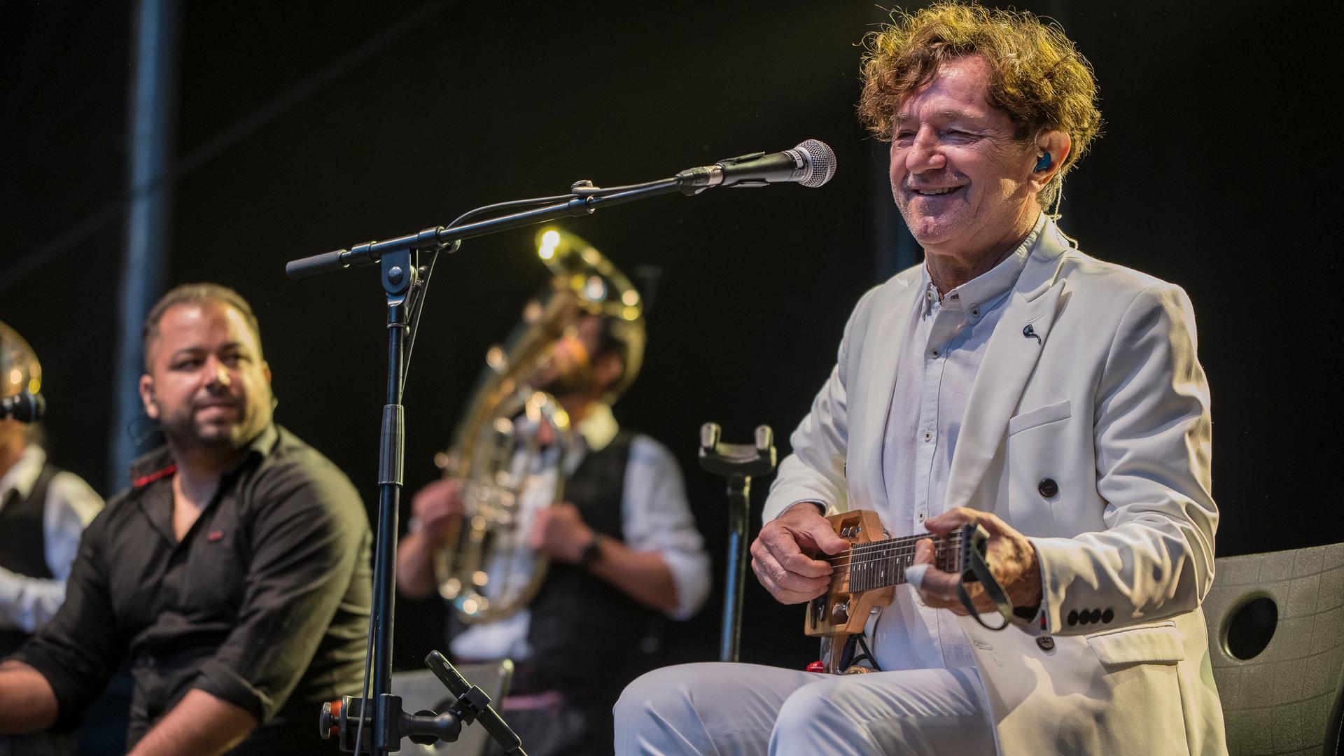 Ein Mann in einem hellen Anzug sitzt auf einem Stuhl, auf dem Schoß hält er eine kleine Gitarre. Vor ihm steht ein Mikrofon. Er hat kurze braune, gelockte Haare und blickt freundlich.