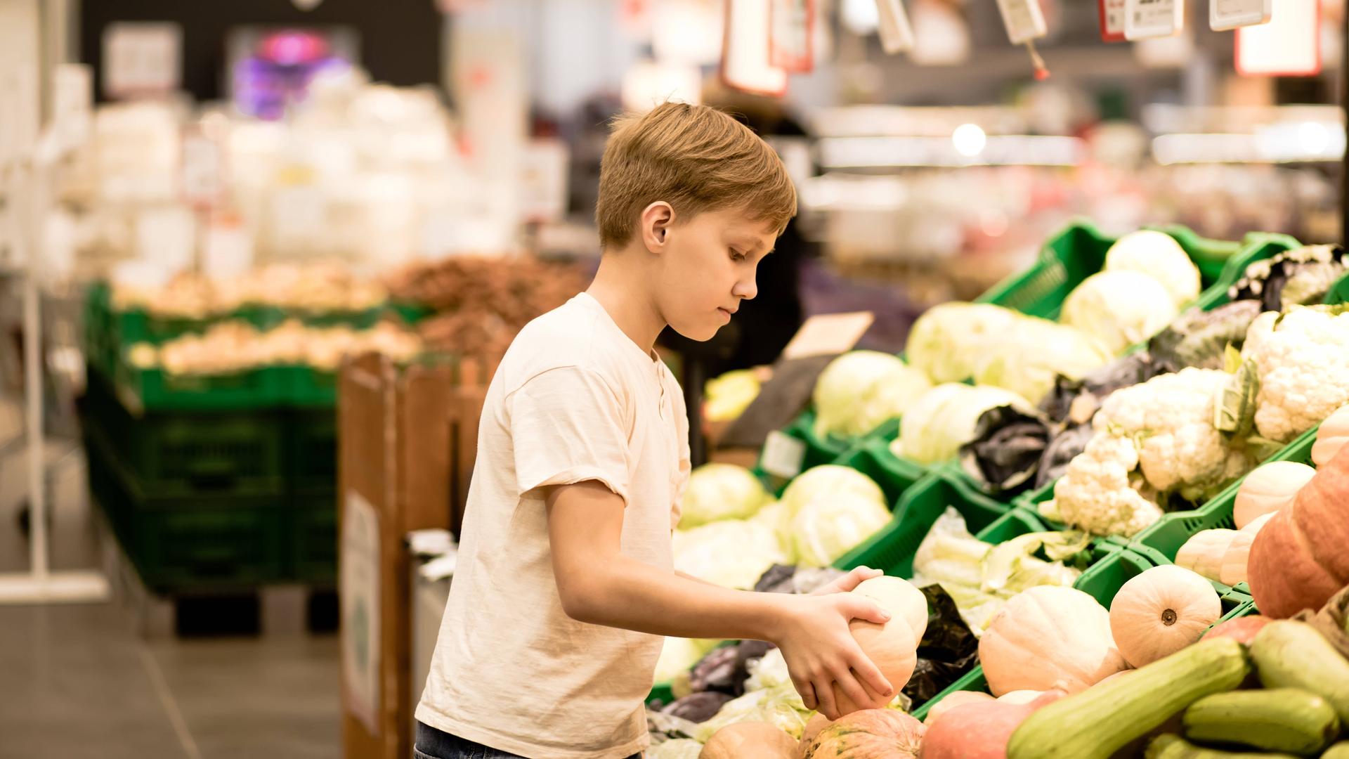 Ein Junge steht beim Gemüse im Supermarkt und hält einen Kürbis in den Händen. Ein Junge steht beim Gemüse im Supermarkt und hält einen Kürbis in den Händen.