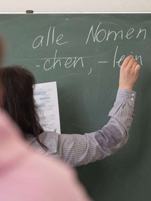 Ein Lehrerin schreibt mit Kreide an einer Tafel, im Vordergrund ist verschwommen ein Schüler von hinten zu sehen