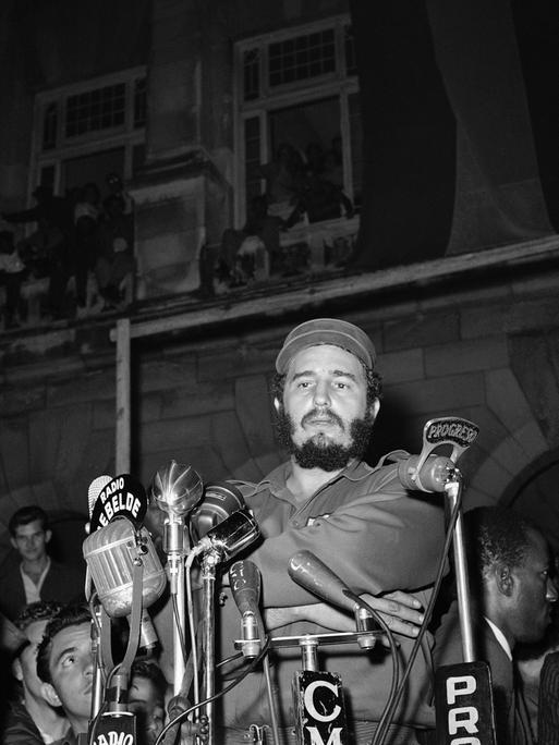 Eine historische scharz-weiß Aufnahme zeigt Fidel Castro angeblitzt vor Mikrofonen am 13. März 1959 vorm Präsidentenpalast in Havanna bei einer Gedenkveranstaltung für die Revolutionäre, die beim Angriff auf den Präsidentenpalast getötet wurden. Der Anschlag wurde von Mitgliedern des Directorio Revolucionario verübt, einer Gruppe, die hauptsächlich aus Universitätsstudenten bestand. Das Directorio arbeitete mit der Bewegung des 26. Juli und anderen revolutionären Gruppen zusammen, um das Batista-Regime zu stürzen.