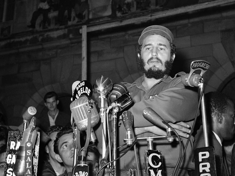Eine historische scharz-weiß Aufnahme zeigt Fidel Castro angeblitzt vor Mikrofonen am 13. März 1959 vorm Präsidentenpalast in Havanna bei einer Gedenkveranstaltung für die Revolutionäre, die beim Angriff auf den Präsidentenpalast getötet wurden. Der Anschlag wurde von Mitgliedern des Directorio Revolucionario verübt, einer Gruppe, die hauptsächlich aus Universitätsstudenten bestand. Das Directorio arbeitete mit der Bewegung des 26. Juli und anderen revolutionären Gruppen zusammen, um das Batista-Regime zu stürzen.
