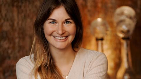 Eine Frau mit dunkelblondem Haar strahlt in die Kamera. In der Hand hält sie Medallien an Bändern. Im warm beleuchteten Hintergrund sieht man weitere Pokale.