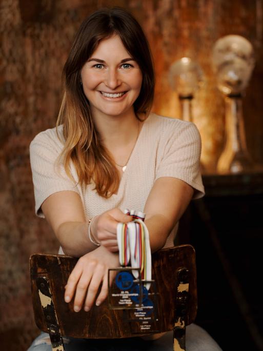 Eine Frau mit dunkelblondem Haar strahlt in die Kamera. In der Hand hält sie Medallien an Bändern. Im warm beleuchteten Hintergrund sieht man weitere Pokale.