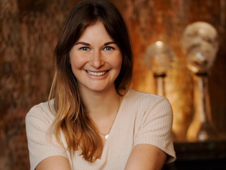 Eine Frau mit dunkelblondem Haar strahlt in die Kamera. In der Hand hält sie Medallien an Bändern. Im warm beleuchteten Hintergrund sieht man weitere Pokale.